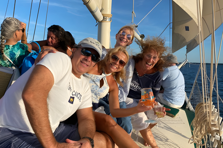 Group of smiling people on a sailboat, enjoying a sunny day with drinks in hand.