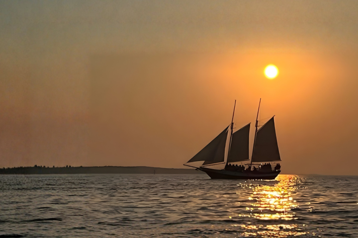 Sailboat on ocean at sunset with orange sky and sun reflection on the water.